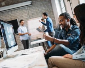 A man and a woman sit at a work desk while gesturing to a desktop computer.